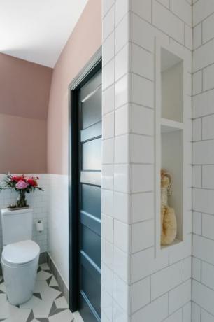 pomalowana na różowo ściana, biała płytka metra, biała toaleta, geometryczne biało-szare płytki, wbudowane regały