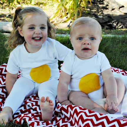 gadis kecil dan bayi duduk di atas selimut berpakaian seperti telur dengan bintik kuning di tengah kemeja putih