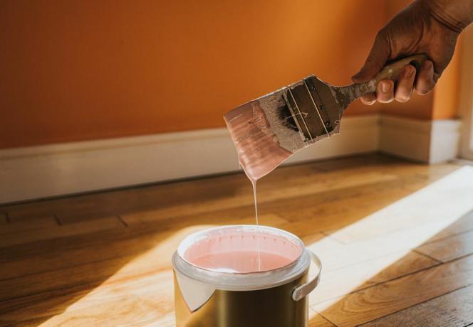 la mano sostiene un pincel grueso y lo quita, después de sumergirlo en la pintura crea una gota de pintura que salpica de vuelta a la maceta, la pared de atrás está pintada de naranja y el color de la lata es rosa, lo que choca conceptualmente con el espacio para Copiar