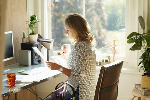 सनी होम ऑफिस में डेस्क पर कॉफी पीती और कागजी कार्रवाई पढ़ती महिला।