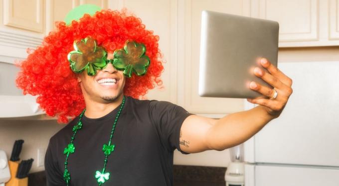 мужчина в рыжем парике и ожерелье дня святого патрика делает селфи на планшете