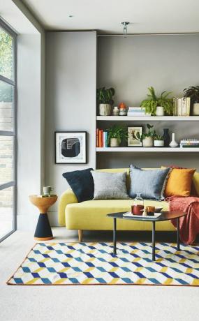 ruang duduk, sofa kuning rak putih di belakang dengan karpet bermotif biru dan kuning di lantai