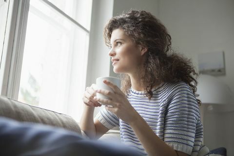 Усмихната млада жена с чаша кафе, гледаща през прозореца