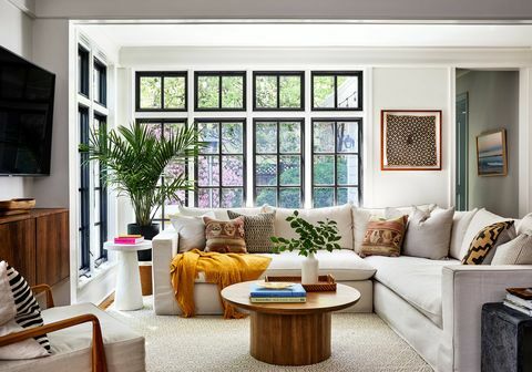 회색 단면 소파, 원형 나무 커피 테이블, 벽걸이 TV, 노란색 깔개 및 장식용 쿠션이 있는 거실