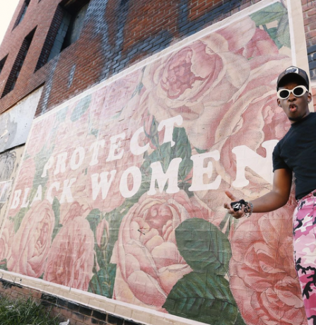seorang pria berdiri di depan mural besar yang bertuliskan " lindungi wanita kulit hitam" di atas latar belakang bunga