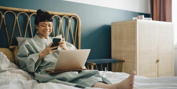 усмихната жена с лаптоп, използваща смарт телефон на легло вкъщи
