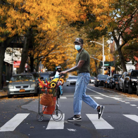 vīrietis, kurš valkā beisbola cepurīti, skatoties kamerā, pāri ielai stumj ratiņus ar ziediem