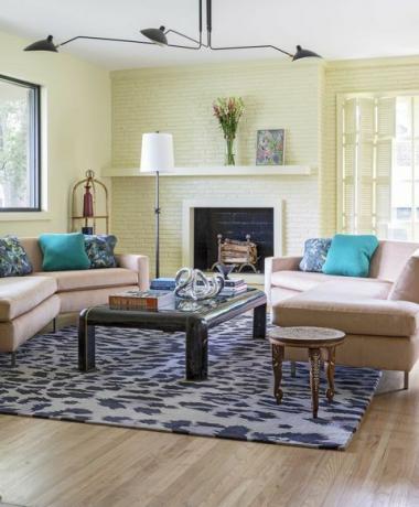 sala de estar con chimenea de ladrillo pintado de amarillo, sofá rosa con gran mesa de café negra y libros de mesa de café, alfombra con estampado de leopardo
