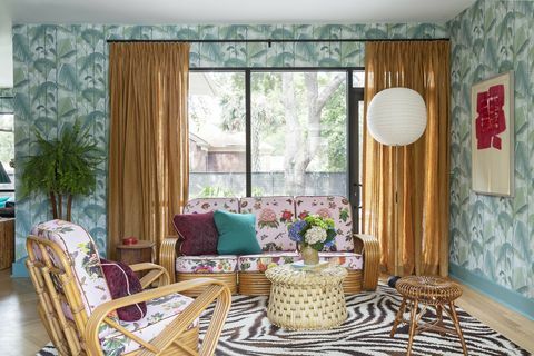 sala de estar con alfombra de área de cebra, papel tapiz de palmeras, cortinas naranjas sobre barras de cortina negras, sofá tropical rosa