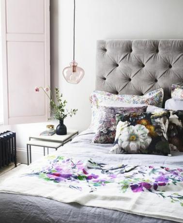 ljust och luftigt sovrum med grå quiltad sängram och blommiga kuddar och rosa fönsterluckor
