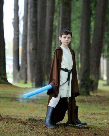 Хлопчик стоїть надворі, одягнений як Обі Ван з блакитною локшиною, як світлова шабля, і довгий коричневий халат