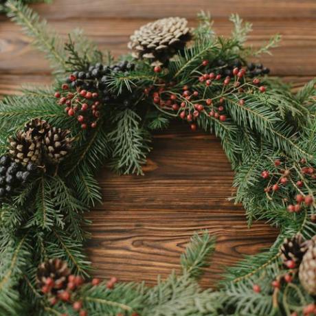 sodoben božični venec z rdečimi jagodami, zelenimi vejami, borovimi storži, okraski iz naravnega drevesa na kmečkem ozadju kmečki božični venec na leseni mizi vesel božič