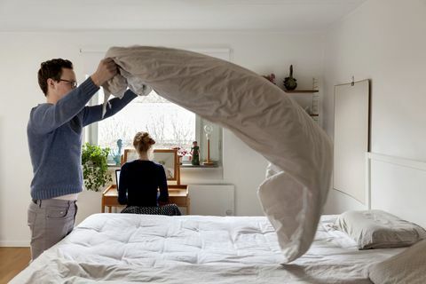 Мъж прави легло, докато жена работи на маса в спалнята