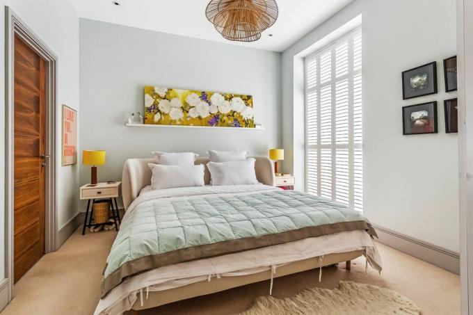 kamar tidur biru pucat dengan tempat tidur, karya seni bunga, lampu gantung pernyataan dan jendela besar dengan tirai rana putih
