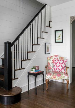 כניסה, עם גרם מדרגות מעץ לבן וכהה, כיסא ישיבה פרחוני צבעוני, שולחן צד שחור