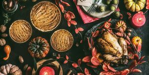 Thanksgiving-Dinner-Tisch mit gebratenem ganzem Huhn oder kleinem Truthahn, Kürbiskuchen auf dunklem rustikalem Hintergrund mit Tellern, Soße, Besteck, Tischdekoration, brennende Kerzen, rote Herbstblätter, Nüsse und festliche Dekoration, Draufsicht, Stillleben, flach legen
