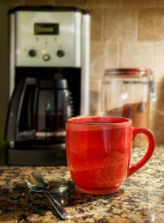 Kouřící se šálek kávy: Červený hrnek vařící kávy spočívá na žulové kuchyňské desce. Na pozadí je vidět kávovar a nádoba na mletou kávu.