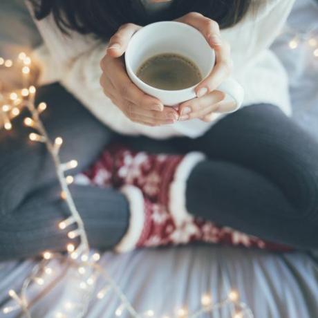 moteris sėdi namuose kojas sukryžiavusi ant lovos, laikydama kavos puodelį, kalėdinę dekoraciją