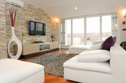 Šiuolaikinė svetainė su vaizdu į balkoną. Yra balta sofa, papuošta dviem didelėmis baltomis pagalvėmis ir viena maža violetine dekoratyvine pagalve. Ant natūralaus akmens sienos visoje sofoje pakabintas plokščiaekranis televizorius. Patalpos centre guli du pufai ir kilimėlis.