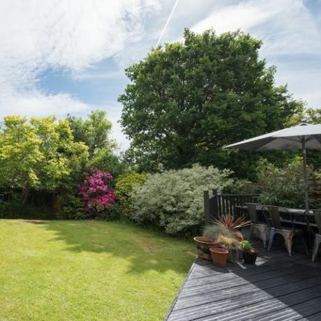 มุมมองทั่วไปของสวนหลังบ้านที่มีพื้นระเบียงสีเทาพร้อมโต๊ะและเก้าอี้ในสวนในวันที่แดดจ้าภายในบ้าน
