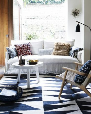 Wohnzimmer, weißes Sofa und blaue Dreiecke Kelim-Teppichfenster hinter einem einfachen Leinensofa und einem weißen Couchtisch sind eine perfekte Kulisse für einen kräftigen blauen und geometrischen weißen Teppich und tonende Kissen
