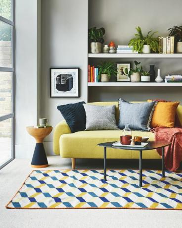 סלון, ספה צהובה מדף לבן מאחור עם שטיח בדוגמת כחול וצהוב על הרצפה