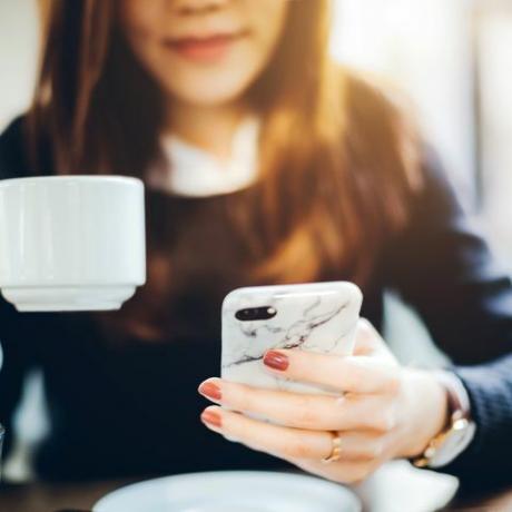 Iš arčiau jaunos moters, anksti ryte prieš darbą besimėgaujančios kava ir skaitančia naujienas mobiliuoju telefonu